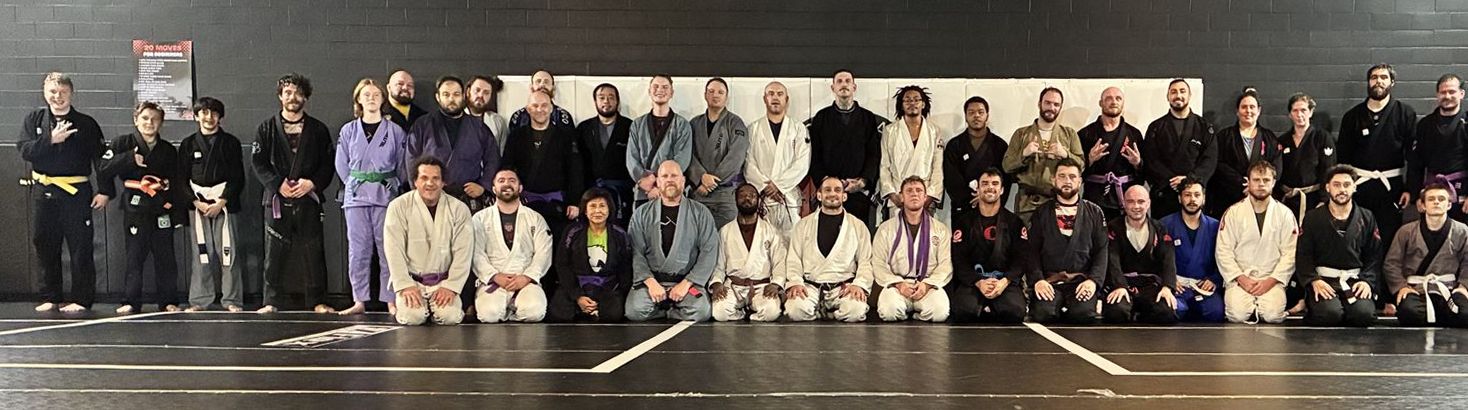 Adult team lined up by belt rank across the academy mats